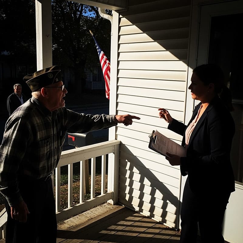 A small American flag displayed on a residential porch, with a stern-looking homeowners association sign visible in the background.